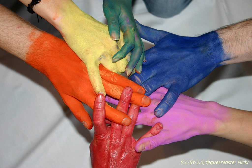 Sechs kreisförmig ineinander verschränkte, mit Regenbogenfarben bemalene Hande
