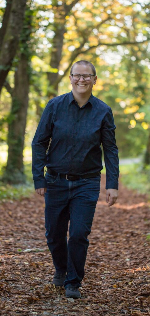 Vero Loitzenbauer, Mitte 30, mit kurz rasierten hellbraunen Haaren, lachendem Gesicht, schwarzer Brille, blauem Hemd und Jean gehend auf Blätterboden in einer Allee