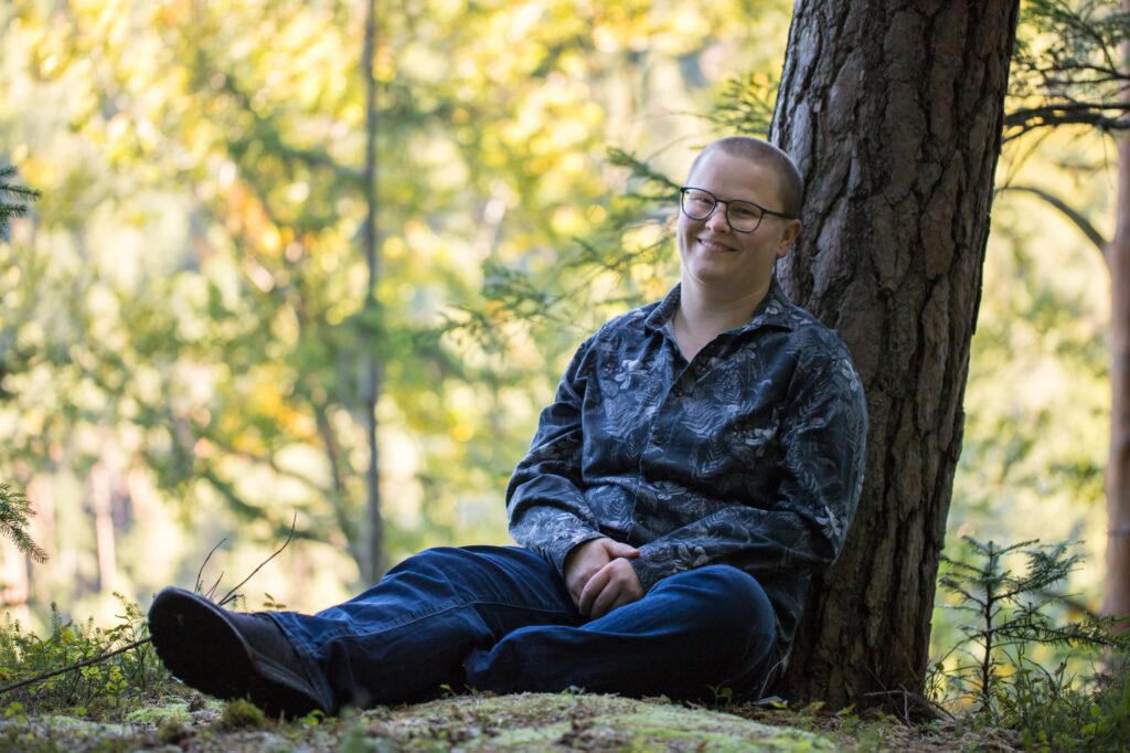 Vero Loitzenbauer entspannt und lächelnd mit schwarz-weißem Hemd mit Pflanzenmuster an einem Baum angelehnt am Waldboden sitzend.